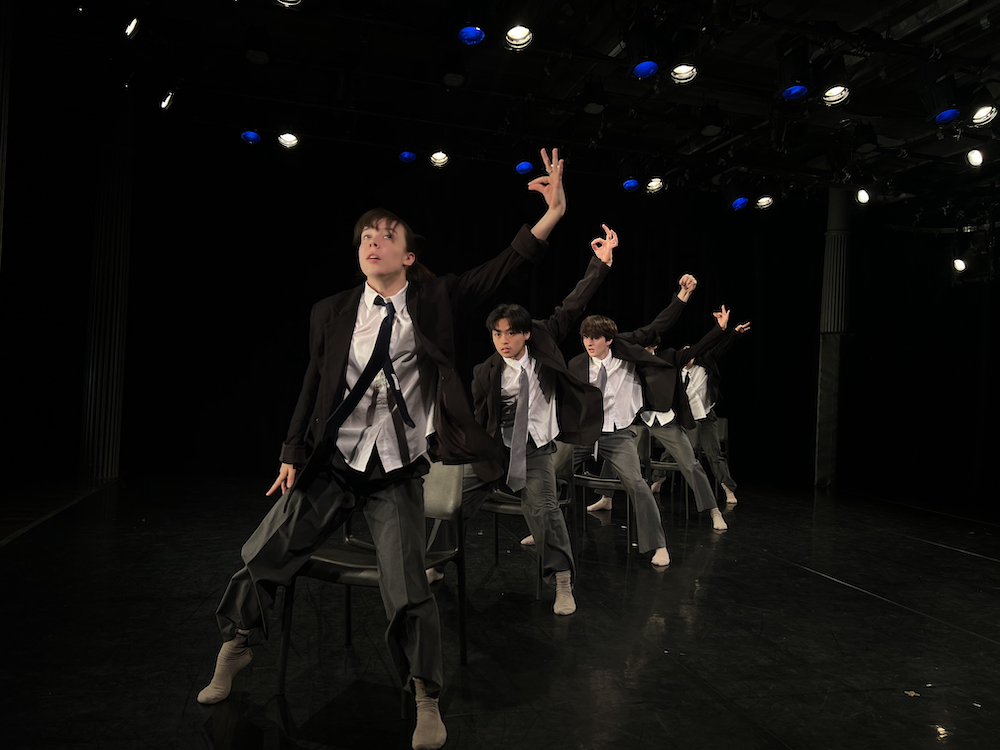 a group of dancers equally dressed in slouchy suits, line up one behind another, rising up off chairs, in unison their left hand poised in an ok signal.